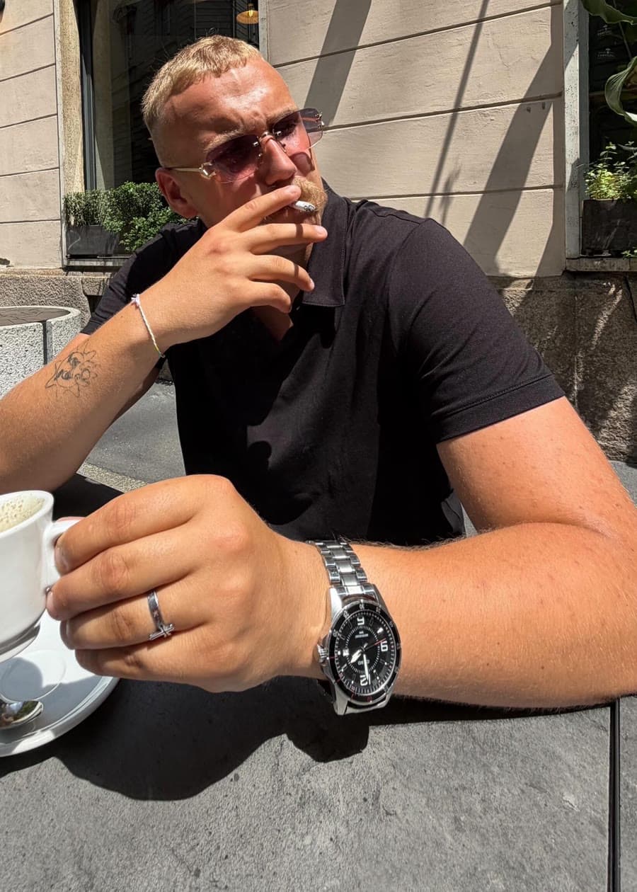 bib$on seated at a café table in a black polo and rose-tinted rimless glasses